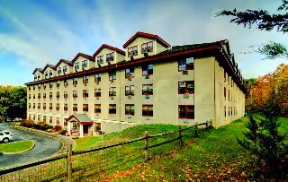 Exterior view of a large multi-story beige building with red roof accents, surrounded by trees and greenery, with a curved driveway and parking area in front.