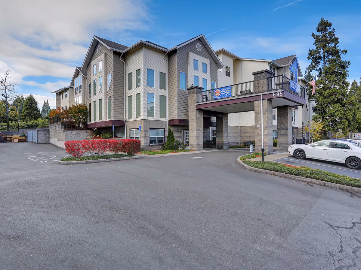 Front exterior of a multi-story senior living building with a covered porte-cochere, landscaping, and parked cars.