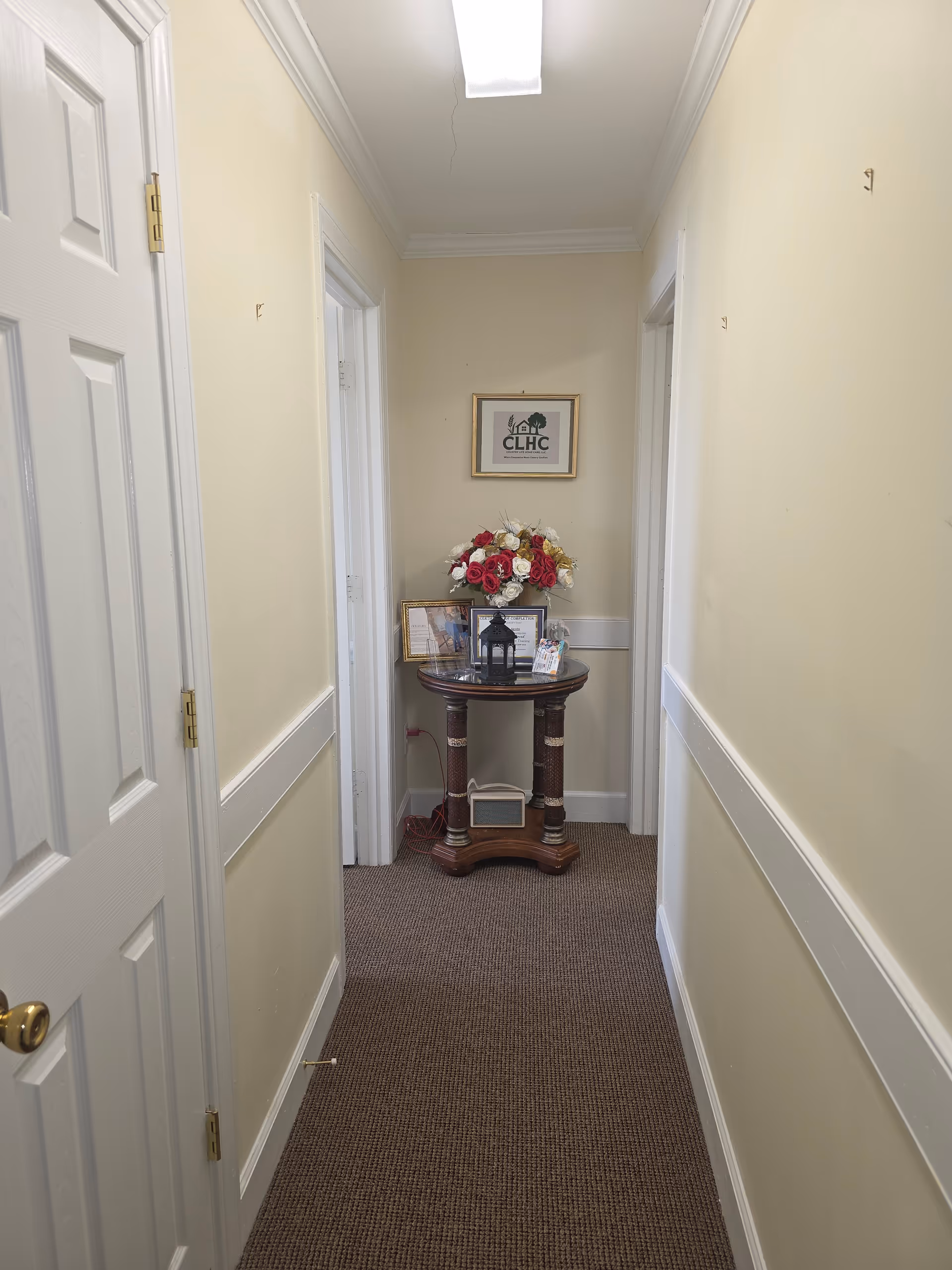 Narrow beige hallway with a wooden pedestal table, flowers, and framed decor.