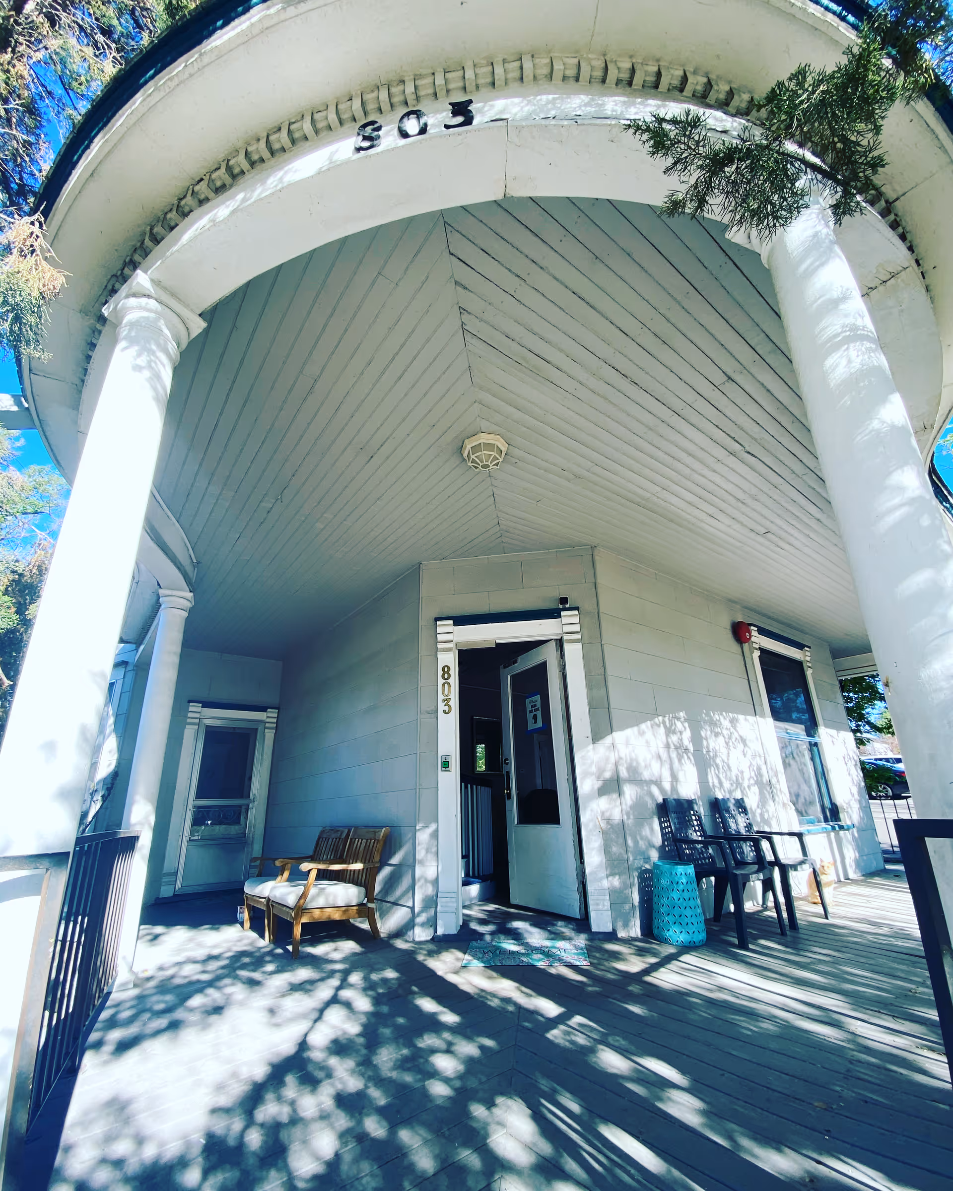 Front porch of a building with white pillars and a curved roof. The building number 803 is displayed above the entrance and on the door frame. There are two cushioned wooden chairs on the left side and two black chairs with a small blue table on the right side of the porch. Shadows of trees are cast on the wooden floor of the porch.