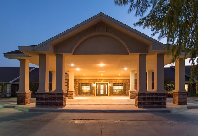 Entrance of a senior living facility named The Rio at Mission Trails during dusk, featuring a covered drop-off area with brick and beige pillars, illuminated by warm lights.