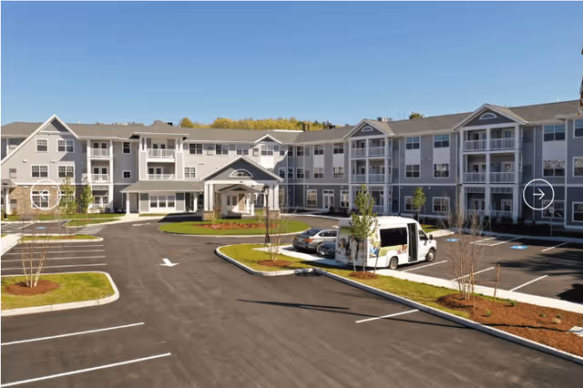 Exterior view of a large, three-story retirement community building with multiple balconies and a covered entrance. The parking lot in front has several parking spaces, a few cars, and a white shuttle van. The sky is clear and blue.