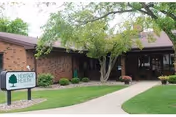 Brick senior living facility exterior with a lawn, trees, and a sign near the entrance.