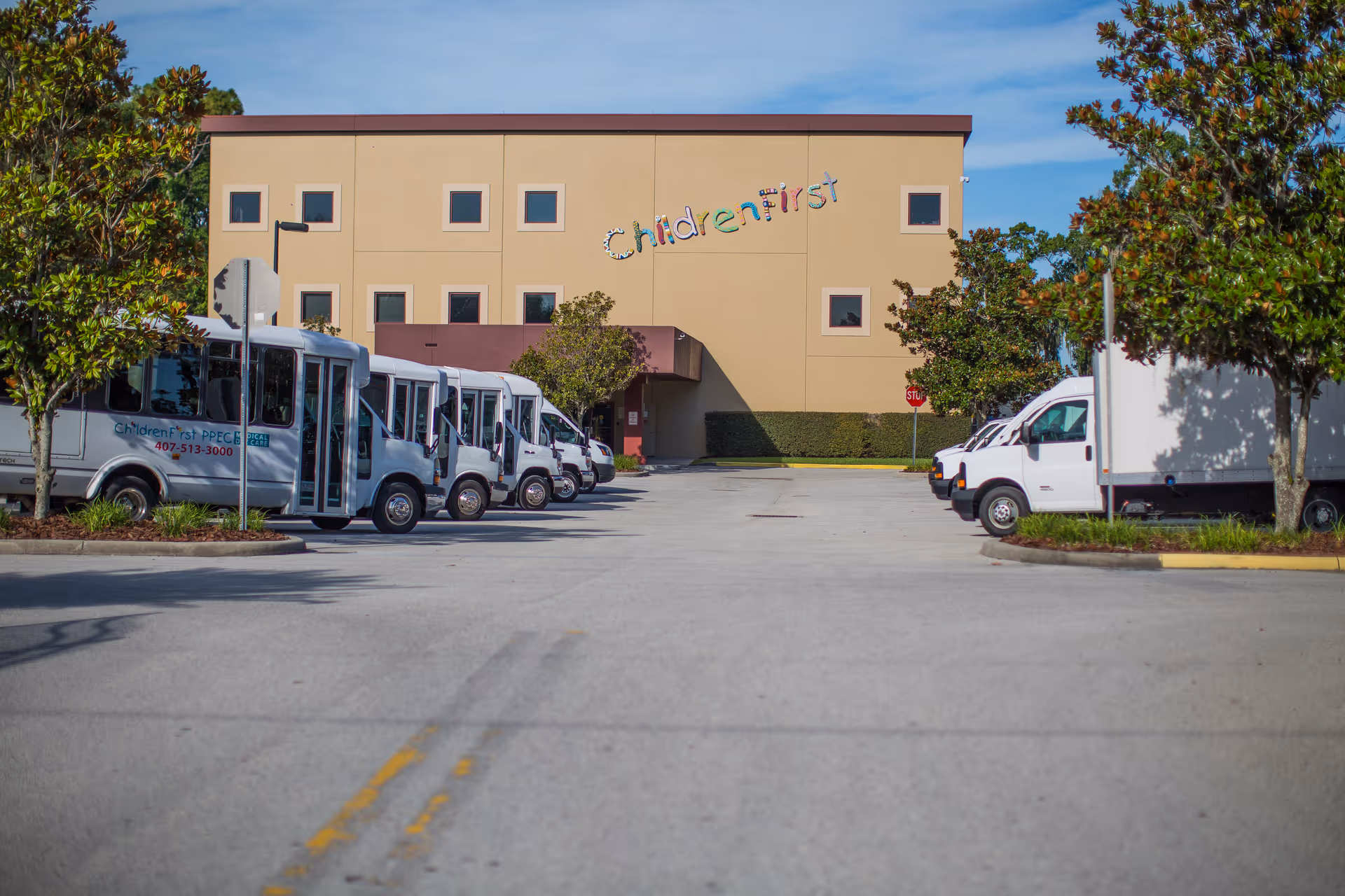 Exterior of a beige building with a colorful sign reading ChildrenFirst and shuttle buses parked nearby.