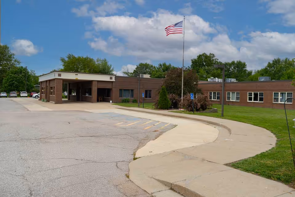 Brick senior care facility with entrance, flagpole, parking, and curved sidewalk.