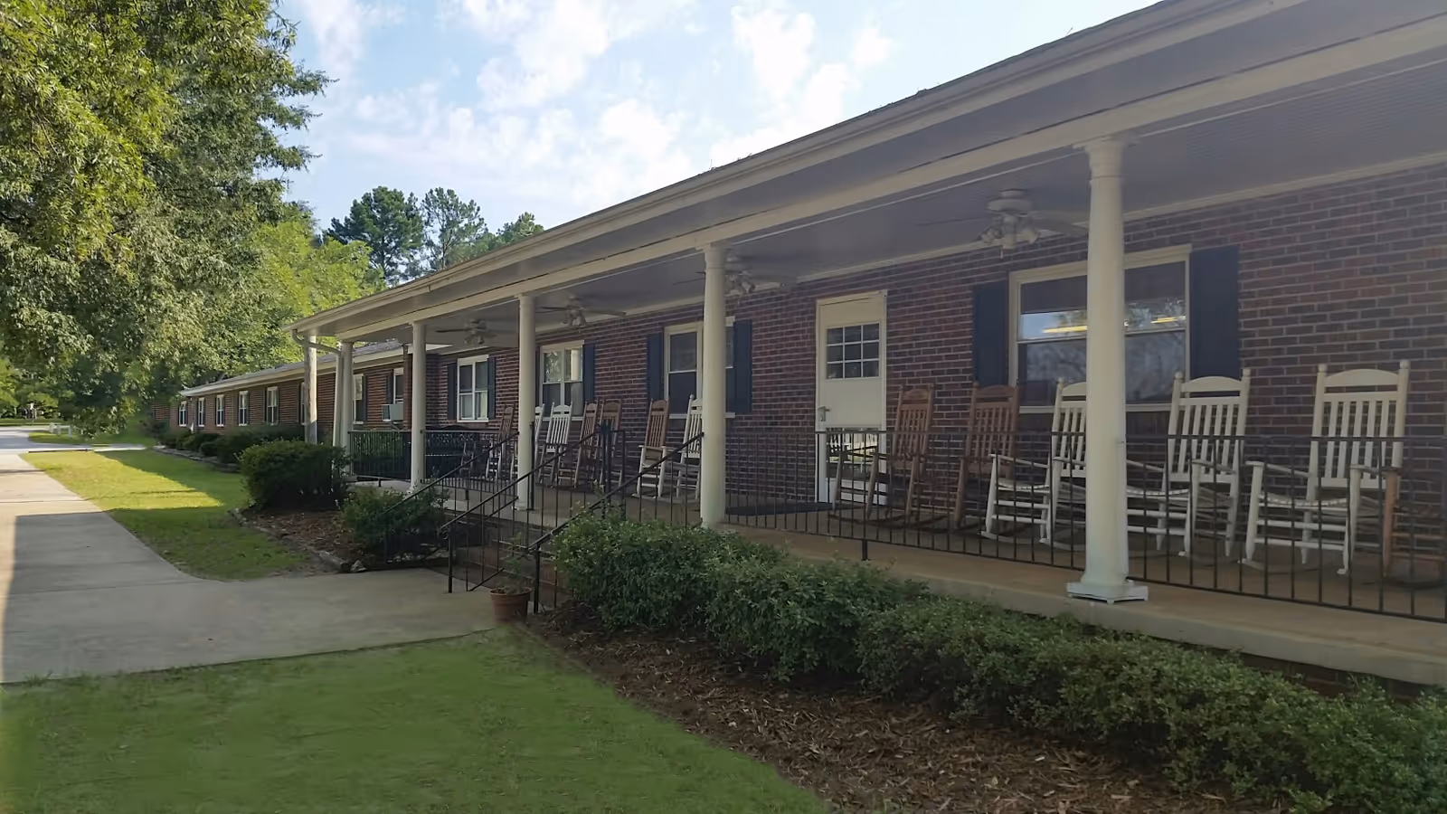 Brick senior living building with a covered porch and rocking chairs along the front.