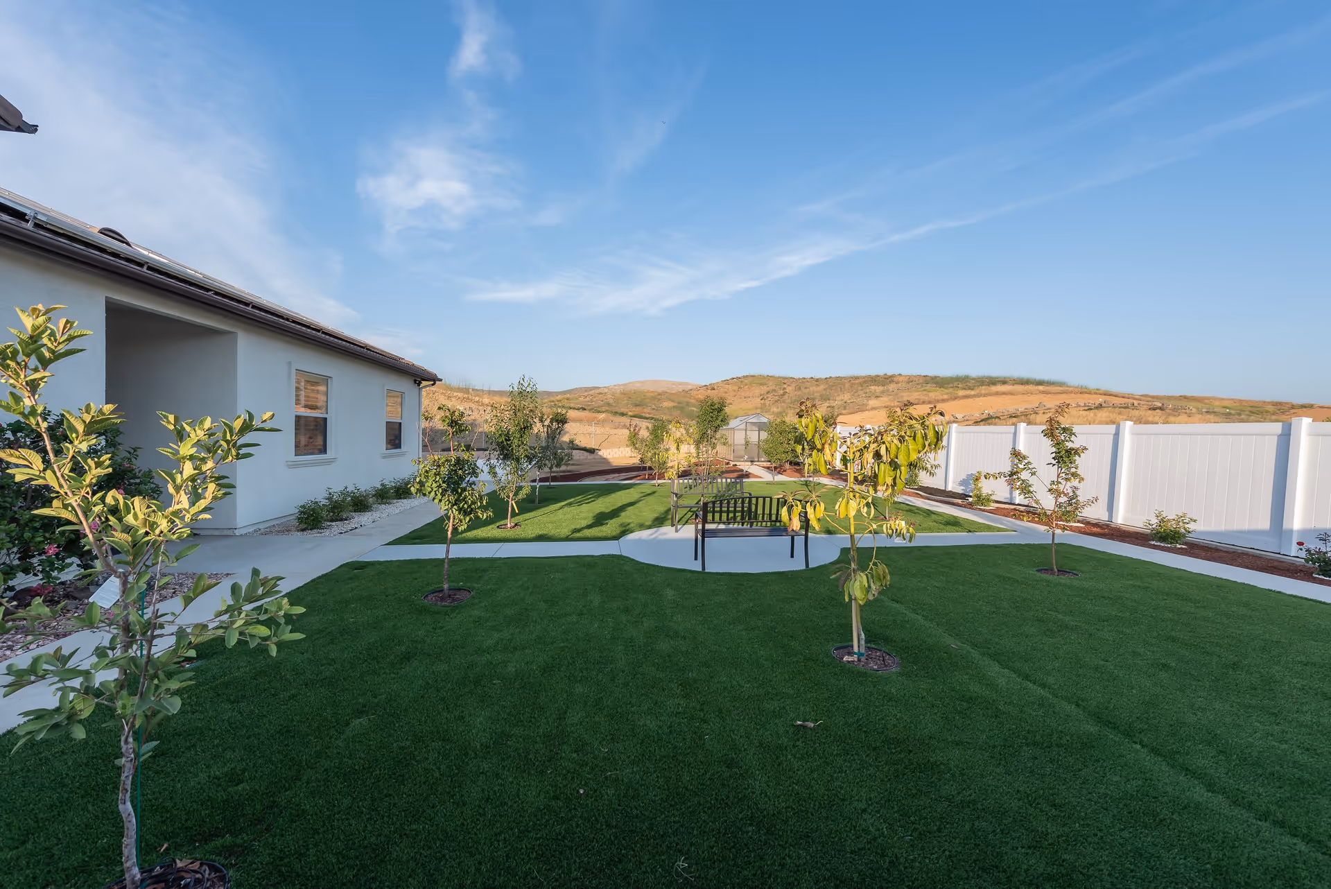 Outdoor garden area at Ohana Excellence Assisted Living featuring a well-maintained lawn with small trees planted throughout. There are paved walkways and a bench seating area in the center. The garden is enclosed by a white fence, with hills visible in the background under a clear blue sky.