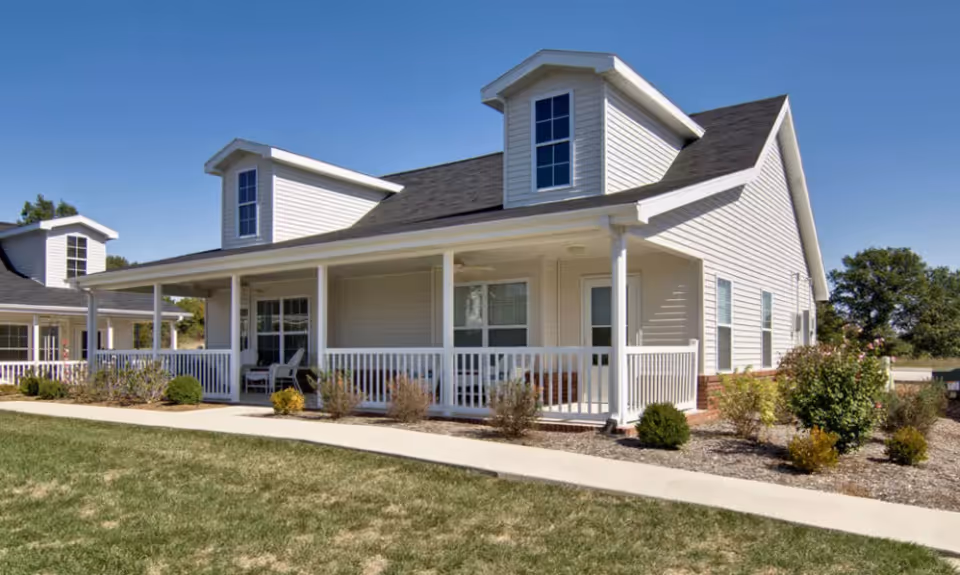 Exterior view of a single-story senior living facility building with white siding, a covered porch with white railings, and dormer windows on the roof. There are shrubs and small bushes planted along the walkway in front of the building under a clear blue sky.