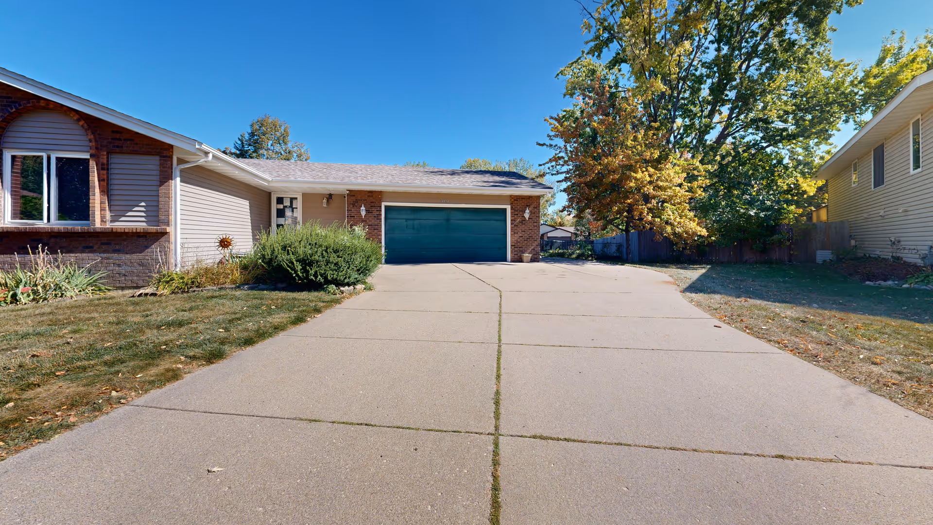 Brick ranch-style house with a long driveway leading to a green garage, trees and blue sky.