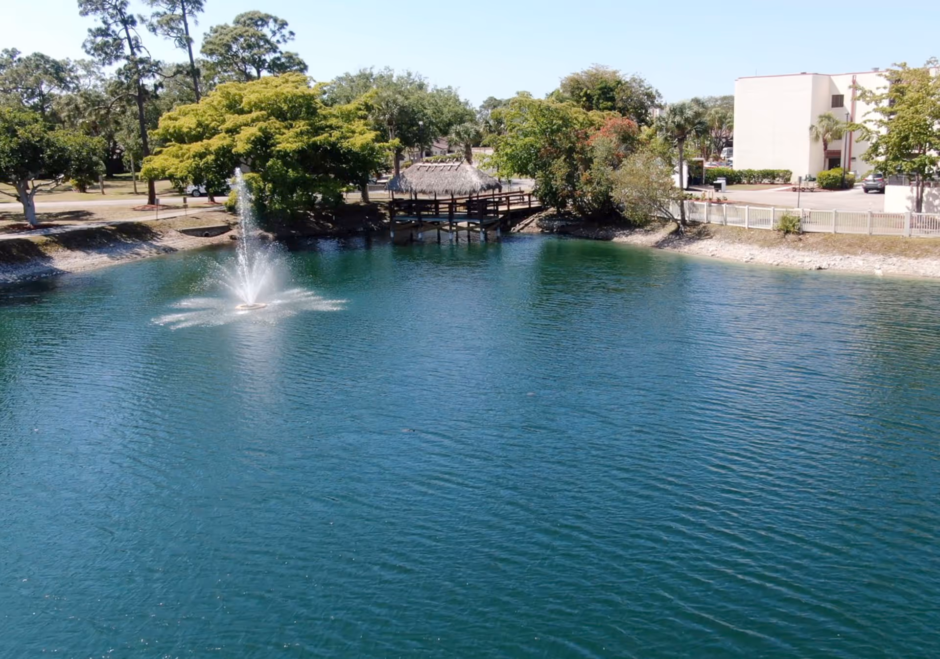 Pond with fountain, trees, a wooden footbridge, and a building in the background.