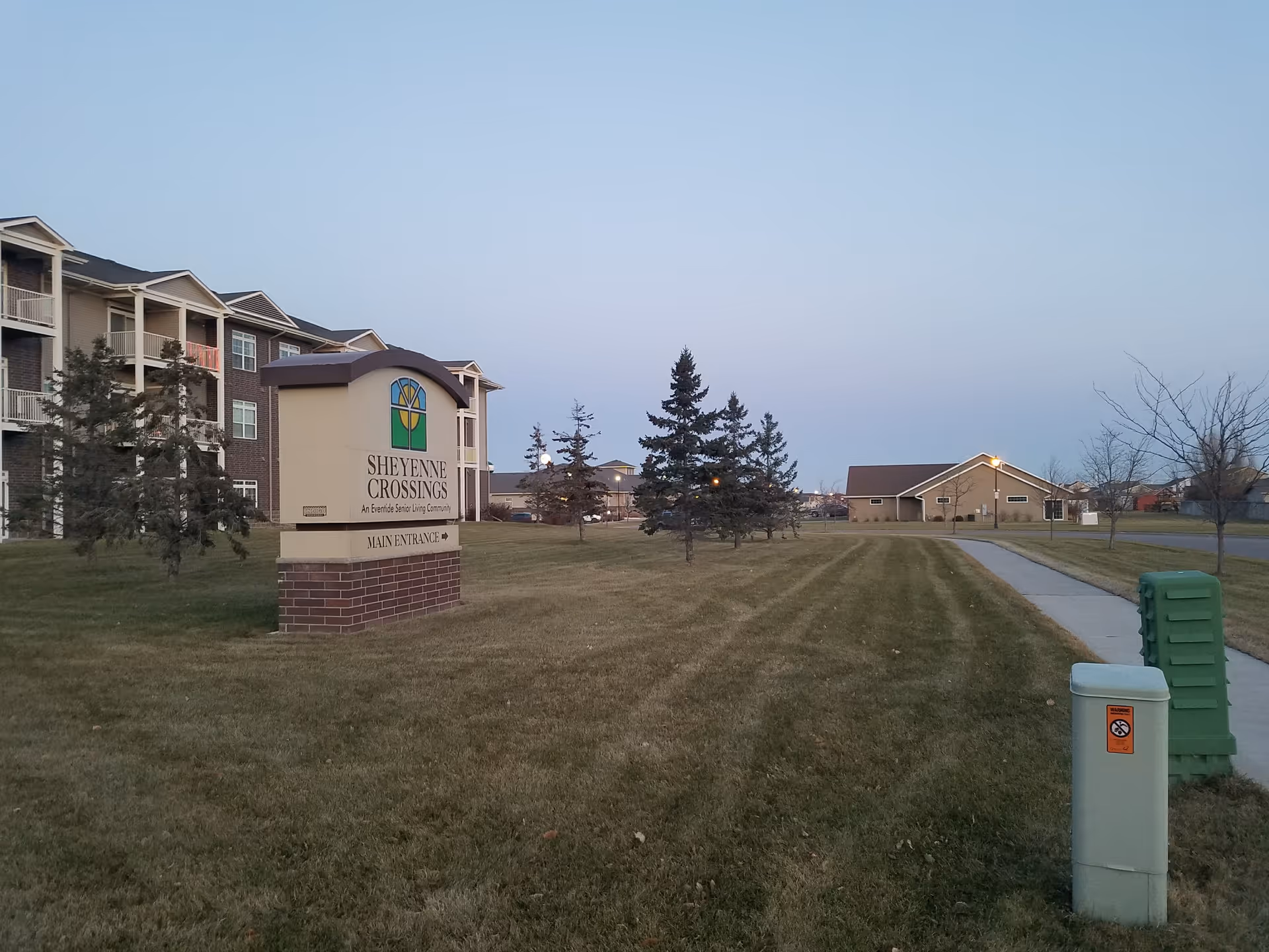 Exterior view of Eventide at Sheyenne Crossings with sign, lawn, trees, and building facade.