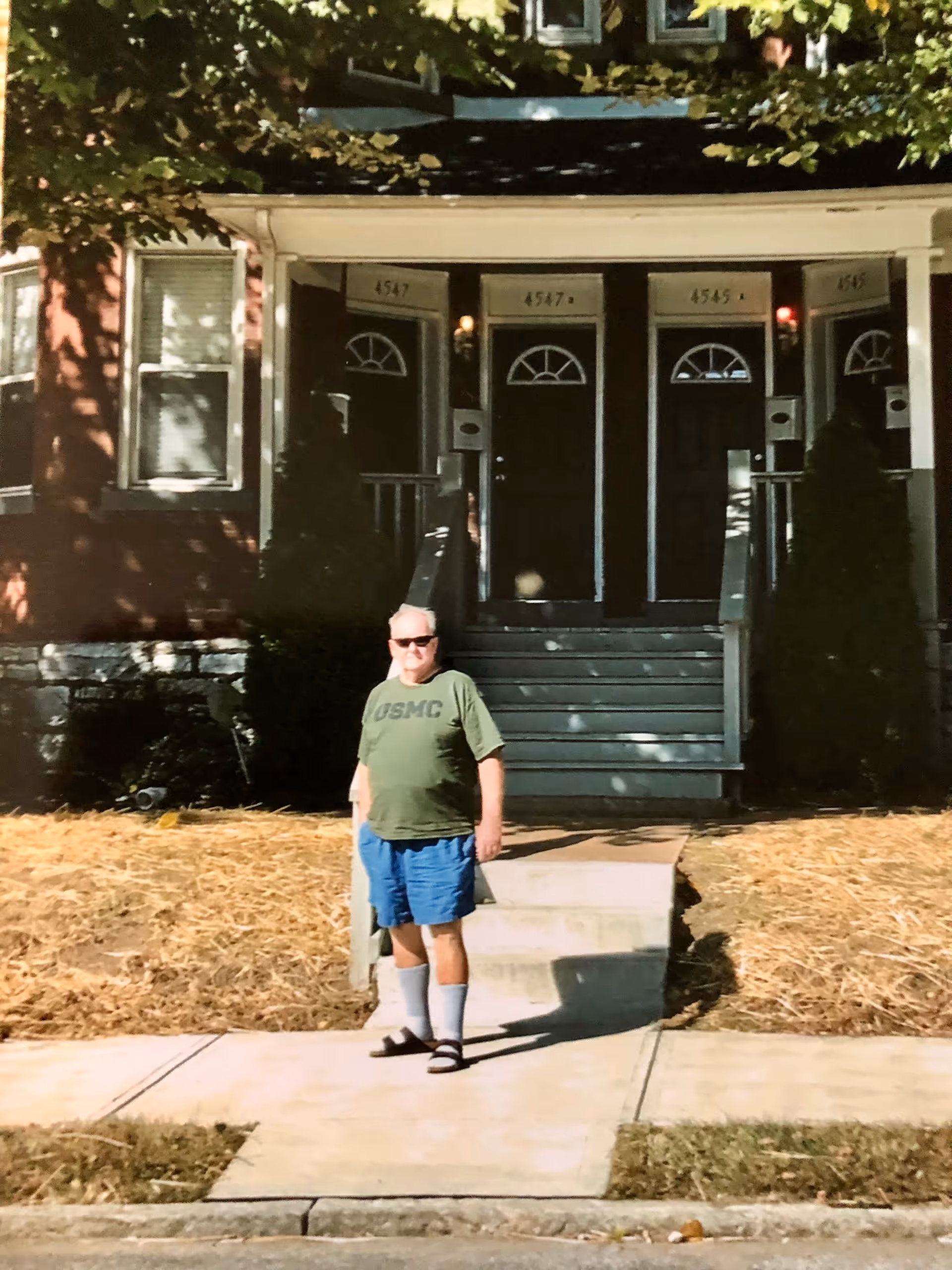 Man wearing green shirt and blue shorts stands on sidewalk in front of row of doors.