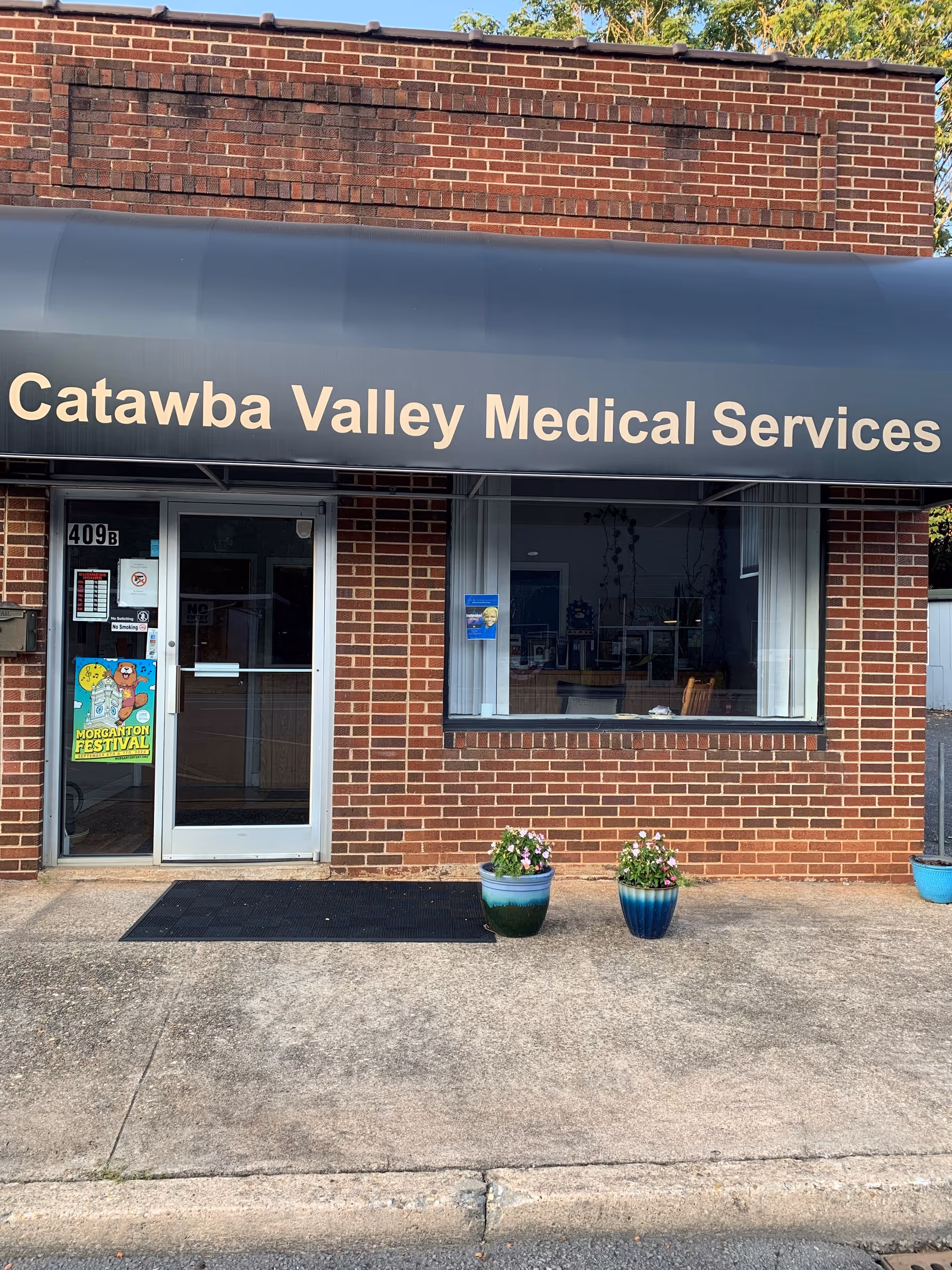 Brick building entrance with a blue awning reading Catawba Valley Medical Services and planters outside.