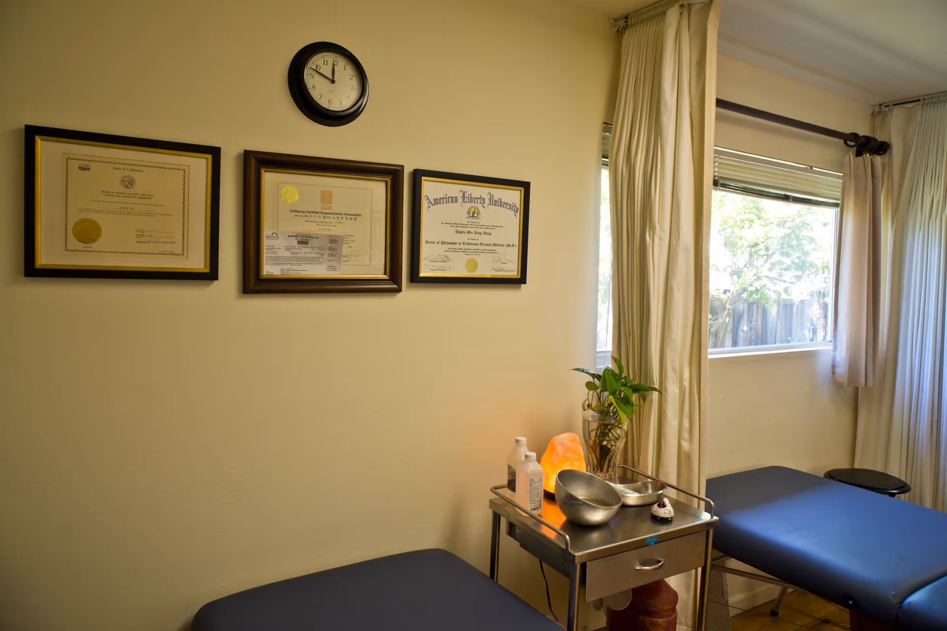 Medical examination room with wall certificates, clock, curtained window, and treatment cart.