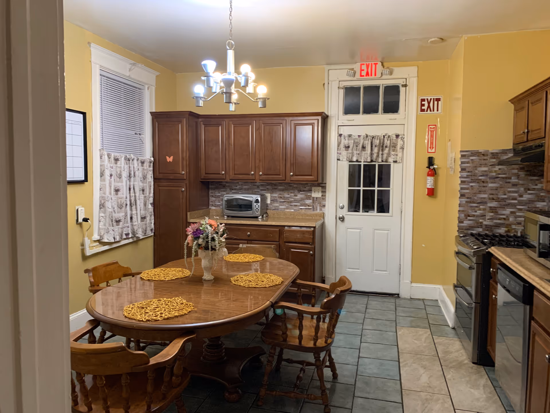 Yellow-walled dining room with a wooden table, chairs, chandelier, and exit door.