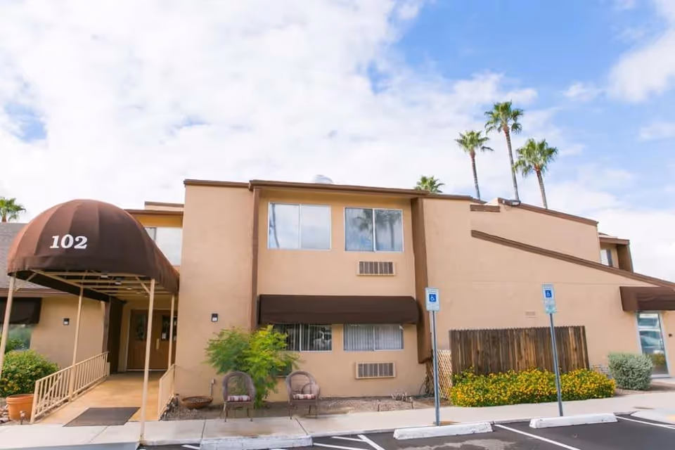 Exterior view of a beige two-story building with a brown awning labeled '102' over the entrance. There are two chairs and a small table near the entrance, some plants and flowers, and three palm trees in the background. The sky is partly cloudy.