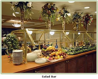 A salad bar with a variety of fresh fruits and vegetables displayed on a wooden counter. There are plates stacked on one side and decorative hanging plants above the bar. The area is well-lit with ceiling lights.