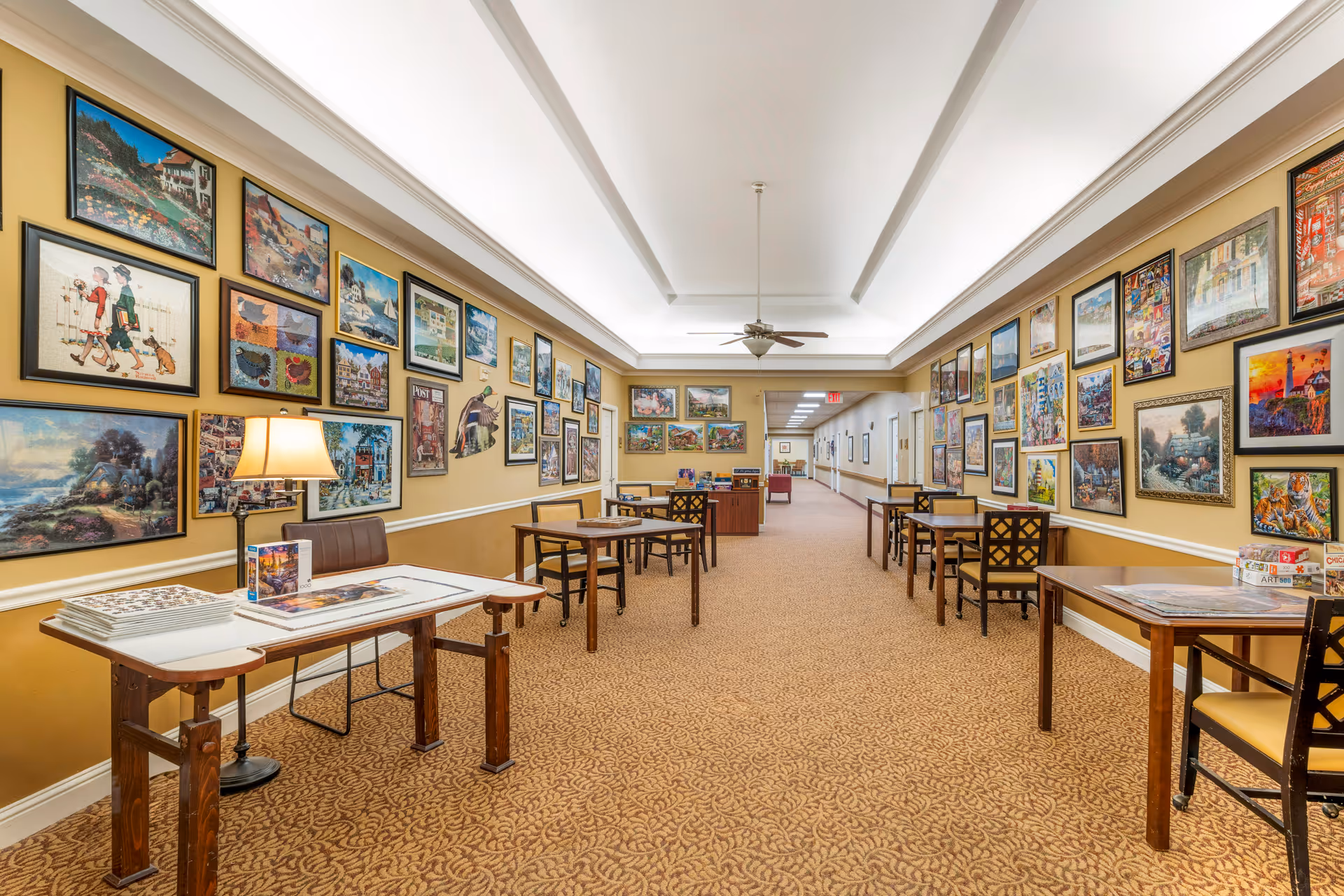 A long hallway in a senior living facility with beige walls adorned with numerous framed pictures and artwork. Several tables and chairs are arranged along the hallway, some with puzzles and books on them. The ceiling has recessed lighting and a ceiling fan, and the floor is carpeted with a patterned design.