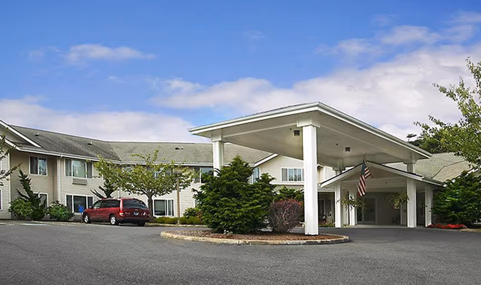 Front exterior of a two-story assisted living building with a covered porte-cochere, landscaping, and a parked red minivan.