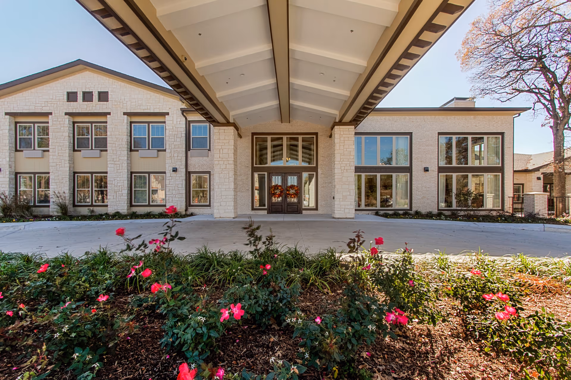 Front exterior view of The Oaks at Flower Mound building with a covered entrance, large windows, and a flower bed with pink flowers in the foreground.