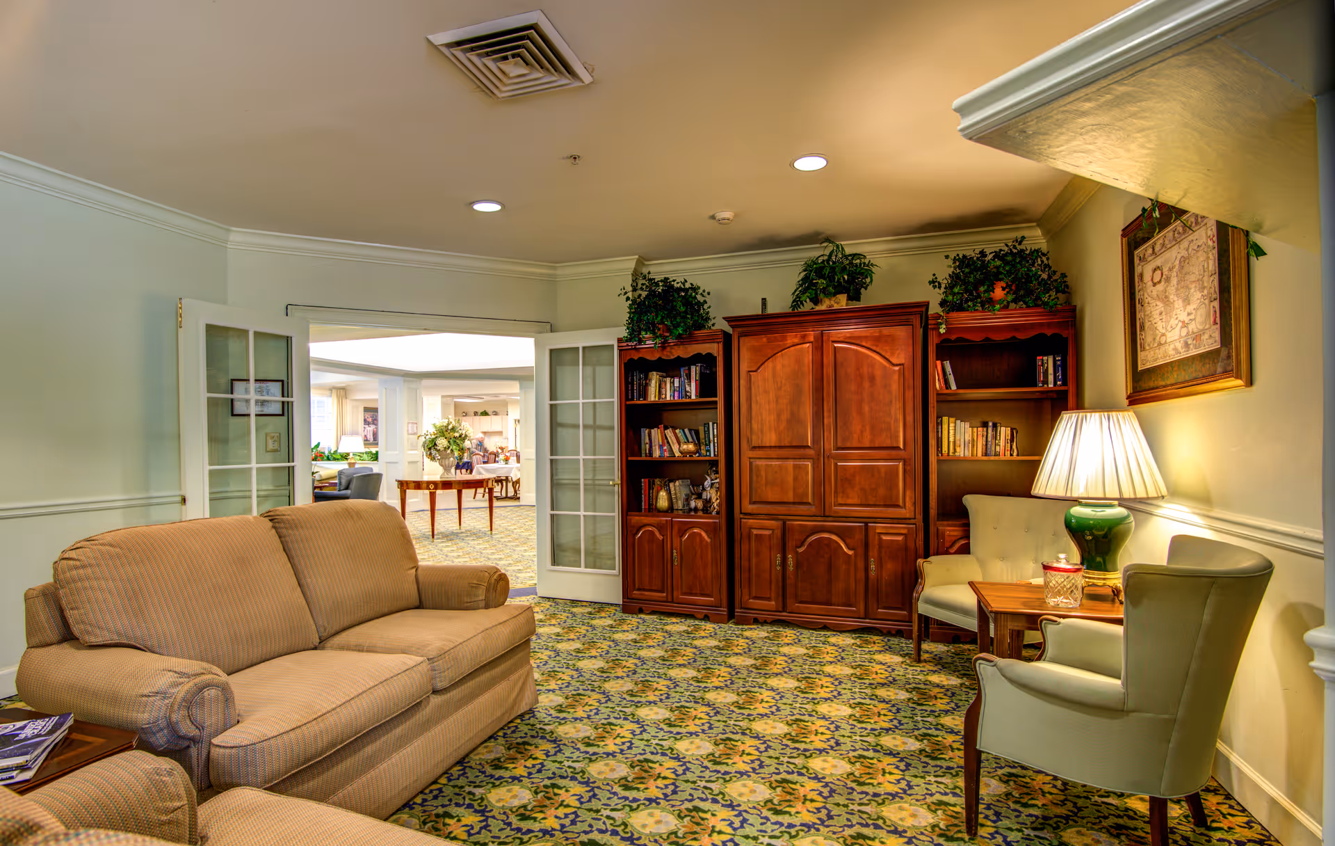 A cozy living room area with a beige sofa, two light green armchairs, a wooden side table with a green lamp, and a large wooden cabinet with bookshelves and plants on top. The room has patterned carpet flooring and light-colored walls. French doors open to another room with more seating and a table with a flower arrangement.