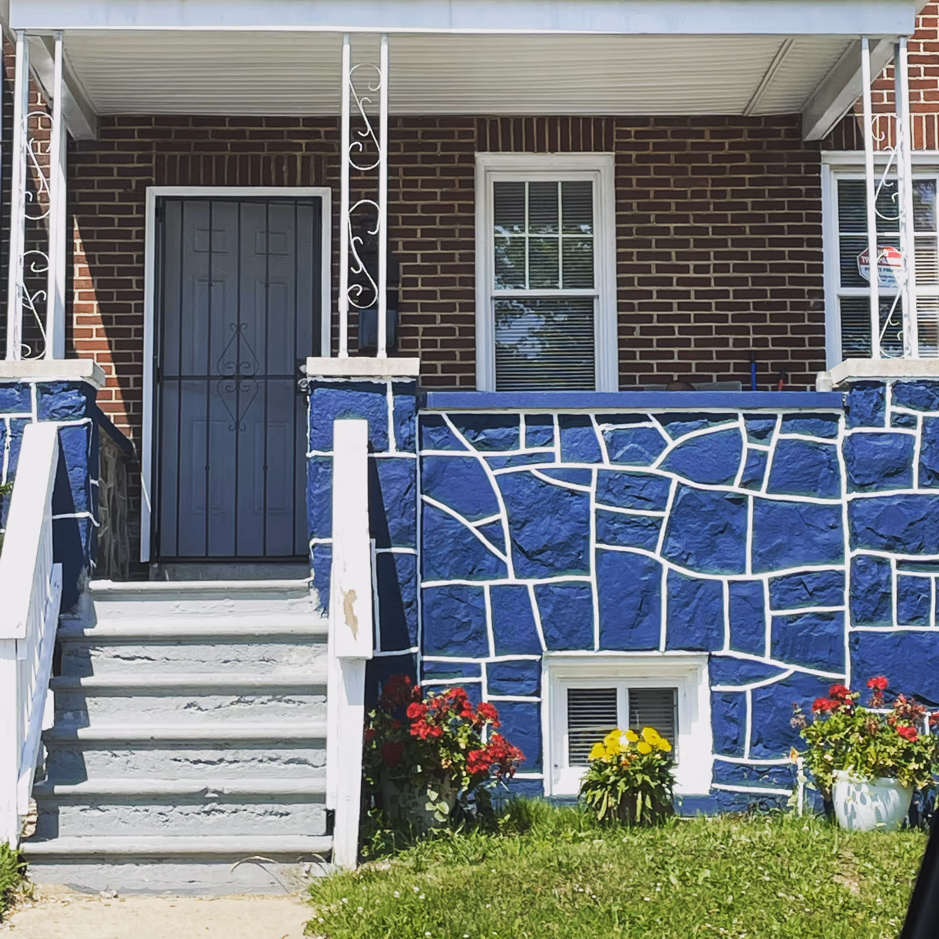Front entrance of a brick house with blue painted stone facing, a gray stairway leading to a metal door, and potted flowers at the base.