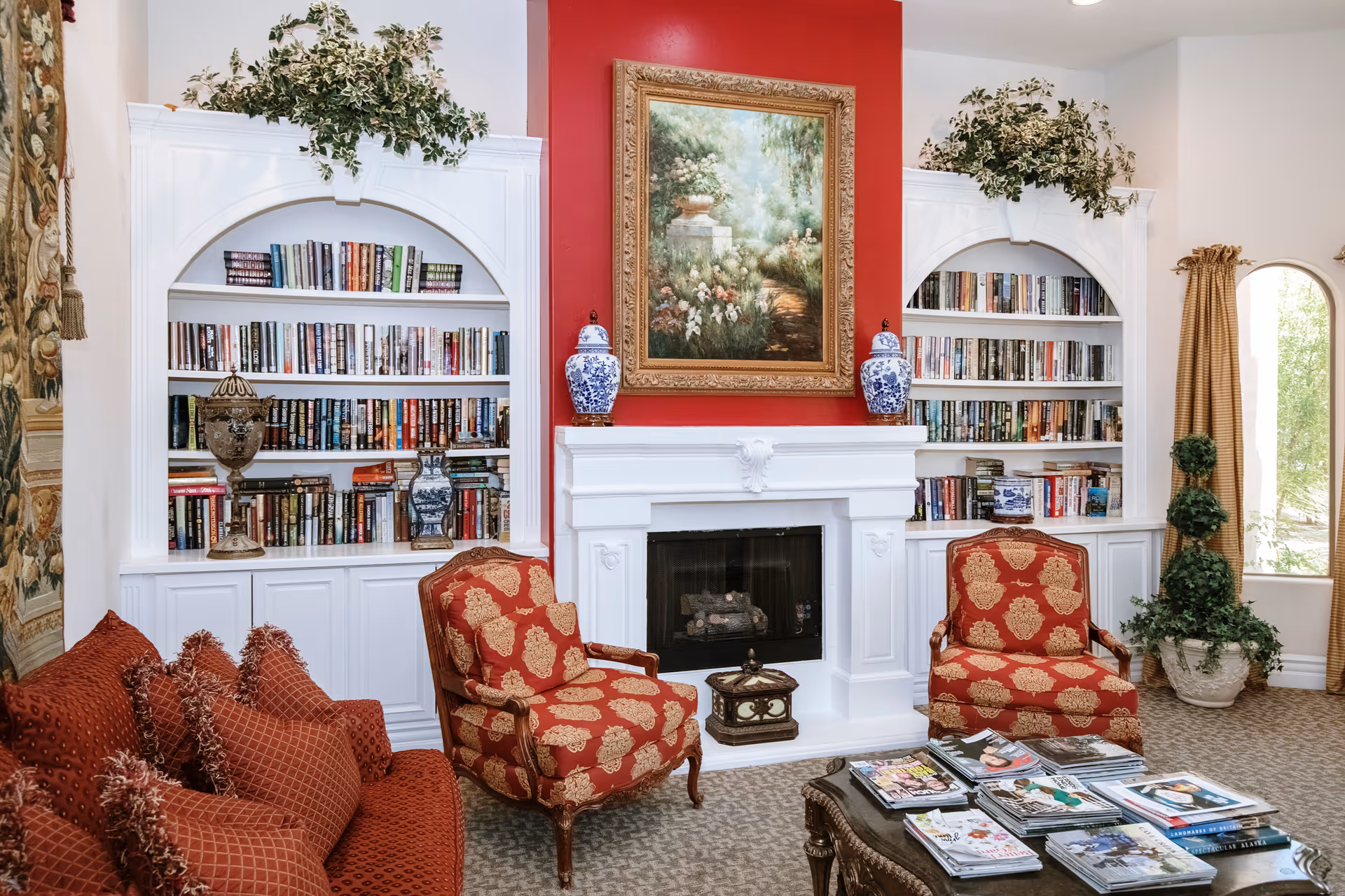 A cozy living room with a white fireplace centered on a red accent wall. Above the fireplace hangs a framed painting of a garden scene. On either side of the fireplace are built-in white bookshelves filled with books and decorative vases. Two red and gold patterned armchairs are placed in front of the fireplace, along with a matching red sofa with multiple cushions. A coffee table in front holds several magazines. Green plants and a window with golden curtains are visible on the right side.