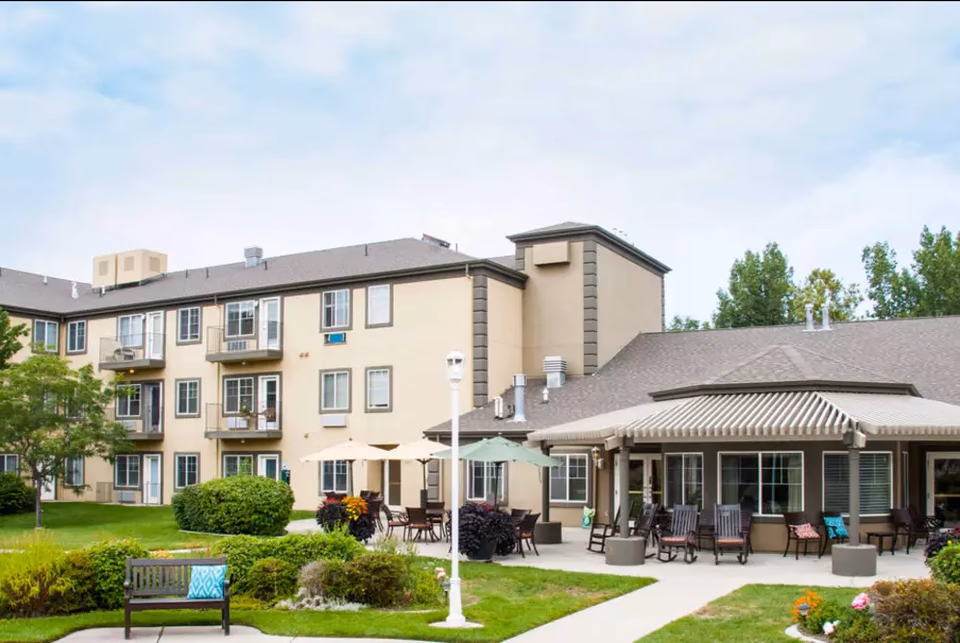 Exterior view of Cedarwood at Sandy senior living facility showing a three-story beige building with balconies, a patio area with tables, chairs, and umbrellas, surrounded by green lawns, bushes, and trees under a partly cloudy sky.