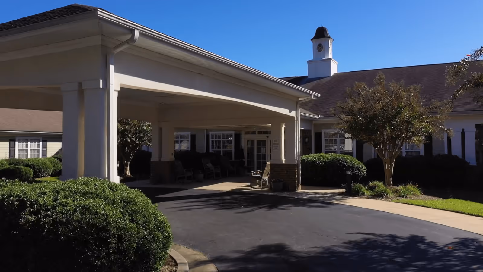 Exterior view of Viva Senior Living at Easley showing the entrance with a covered driveway, surrounded by bushes and trees under a clear blue sky.