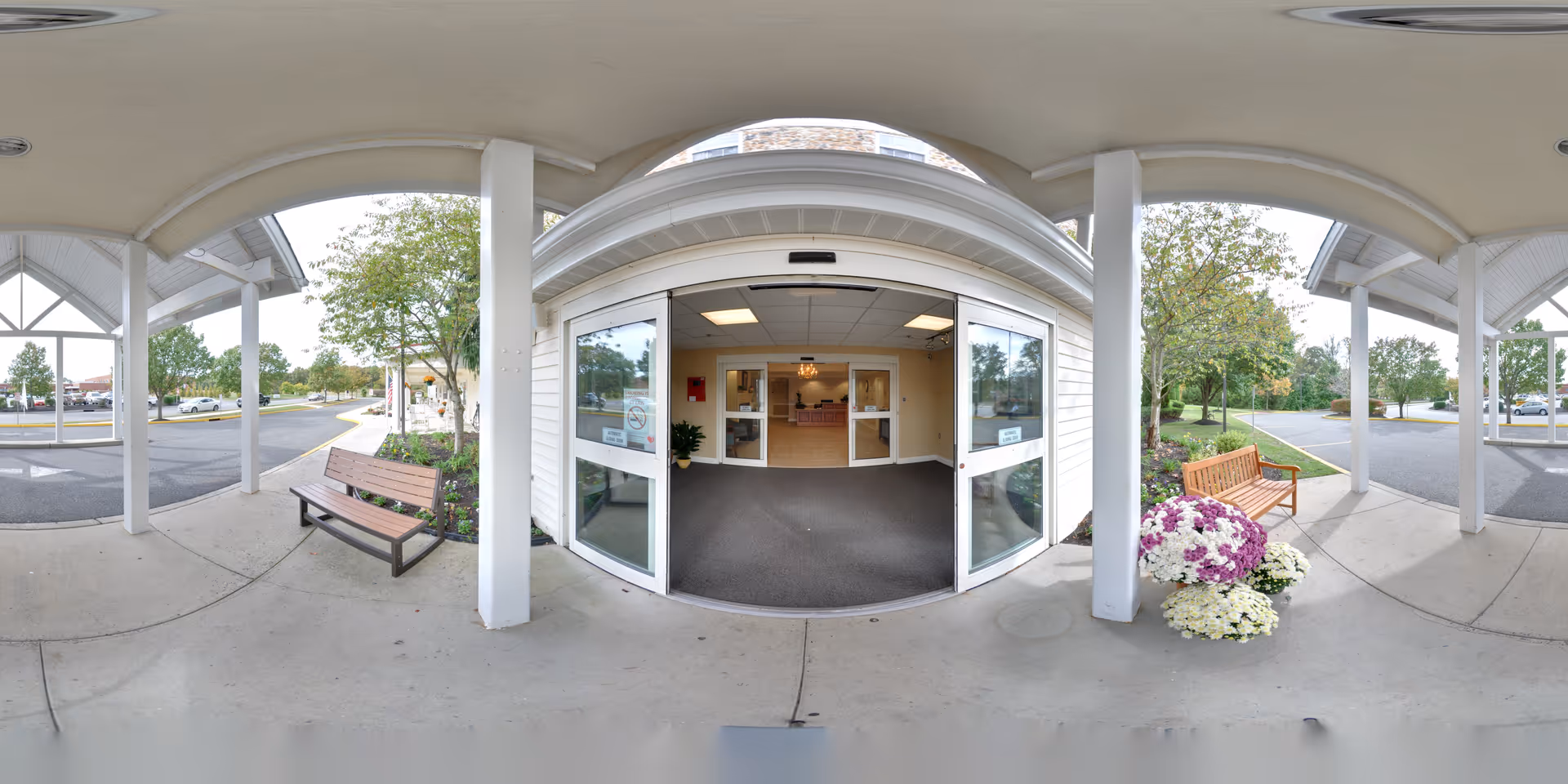 Covered entrance area of Washington Township Senior Living with automatic sliding glass doors leading inside. There are benches on either side of the entrance and flower arrangements near one bench. Trees and parking areas are visible in the background.