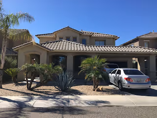 Two-story beige assisted living home with tiled roof, palms, and a white car in driveway.