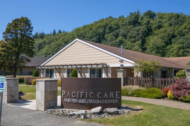 Exterior front of Pacific Care and Rehabilitation Center with a large sign, landscaped grounds, and wooded hills in the background.