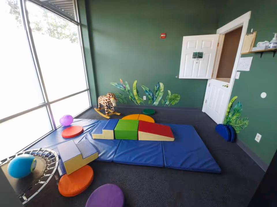 Bright interior therapy room with colorful foam mats, soft shapes, and a plush giraffe toy.