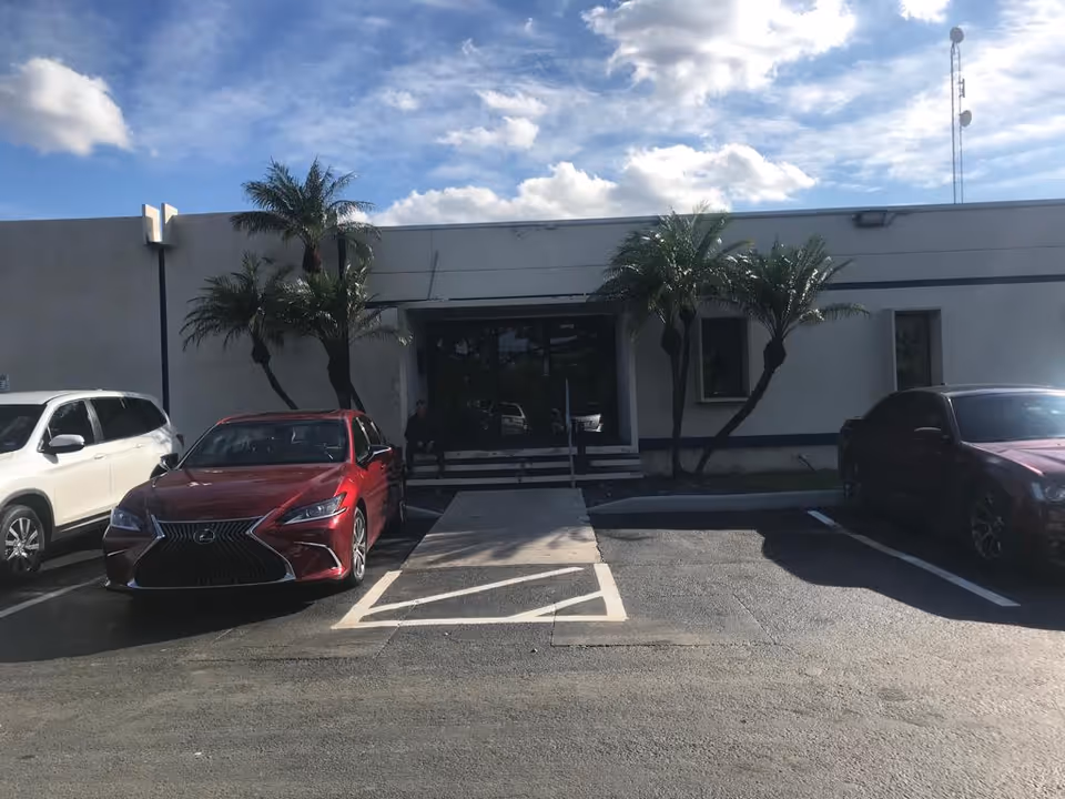 Front view of a single-story facility entrance with parked cars and palm trees.