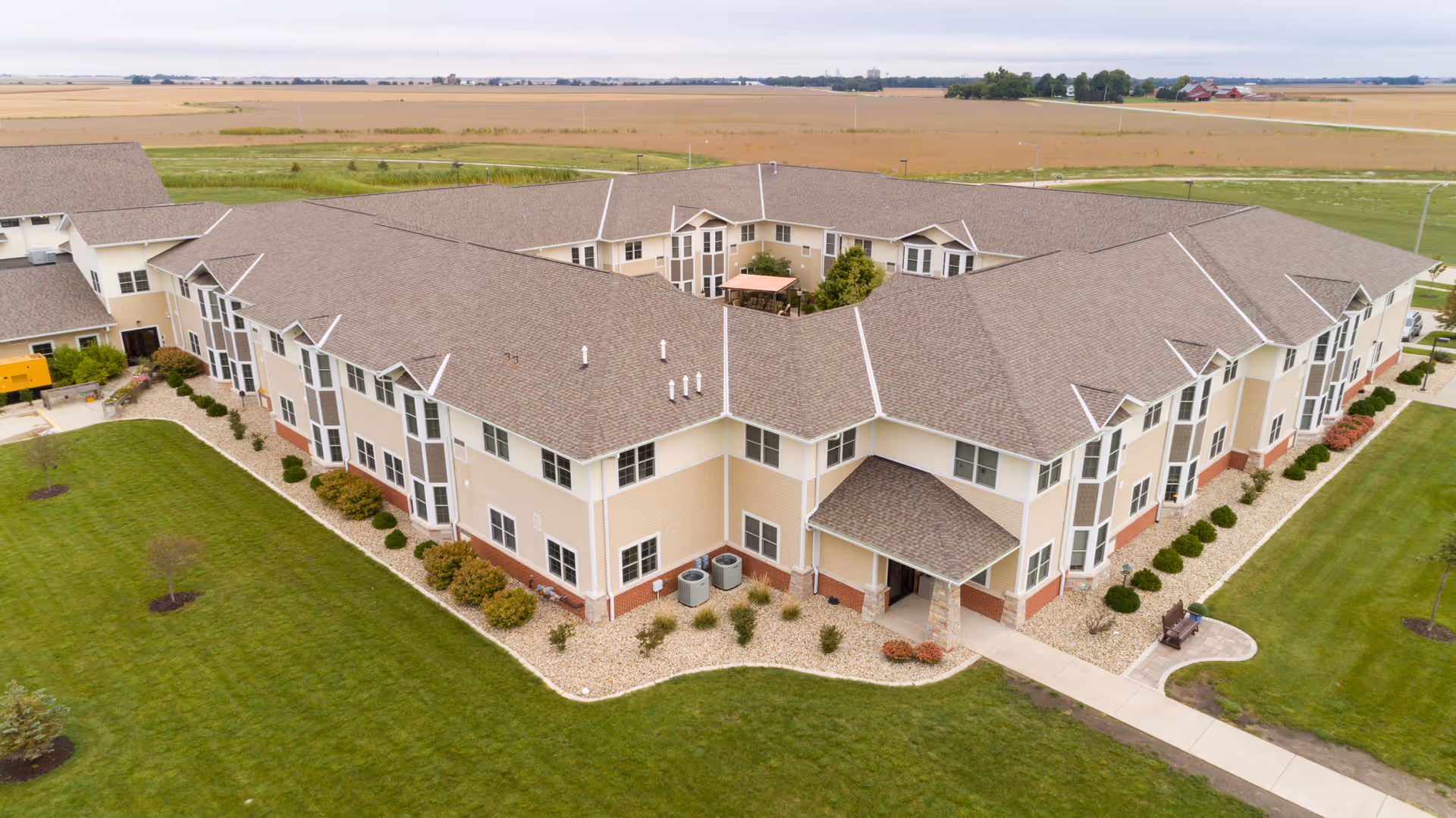 Aerial view of a large, two-story senior living facility building with beige and white exterior walls and a brown shingled roof. The building is surrounded by well-maintained green lawns, landscaped bushes, and a paved walkway leading to the entrance. In the background, there are open fields and a few distant structures under a cloudy sky.