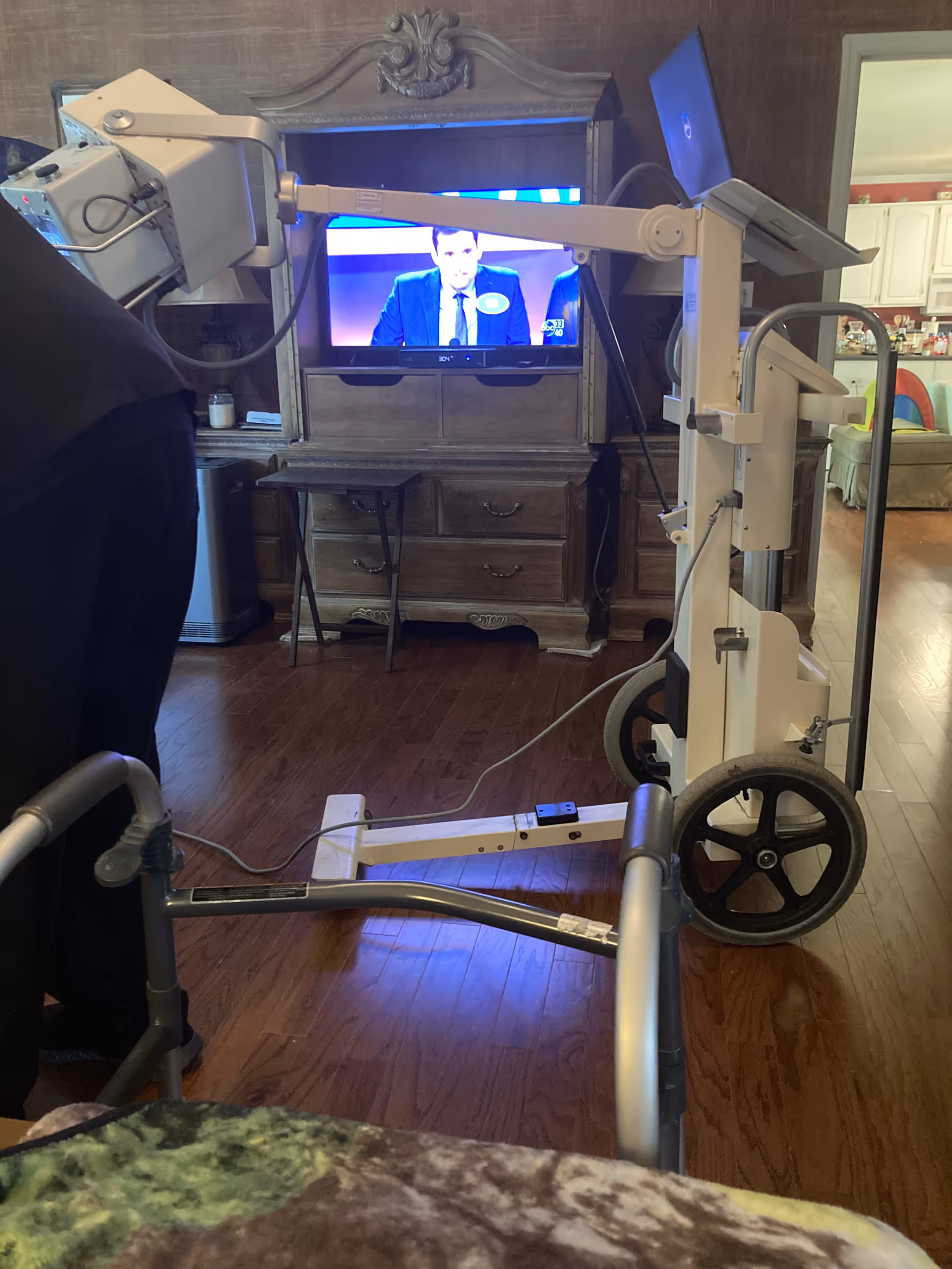 A wheeled medical device stands in a living room with a TV and ornate cabinet.