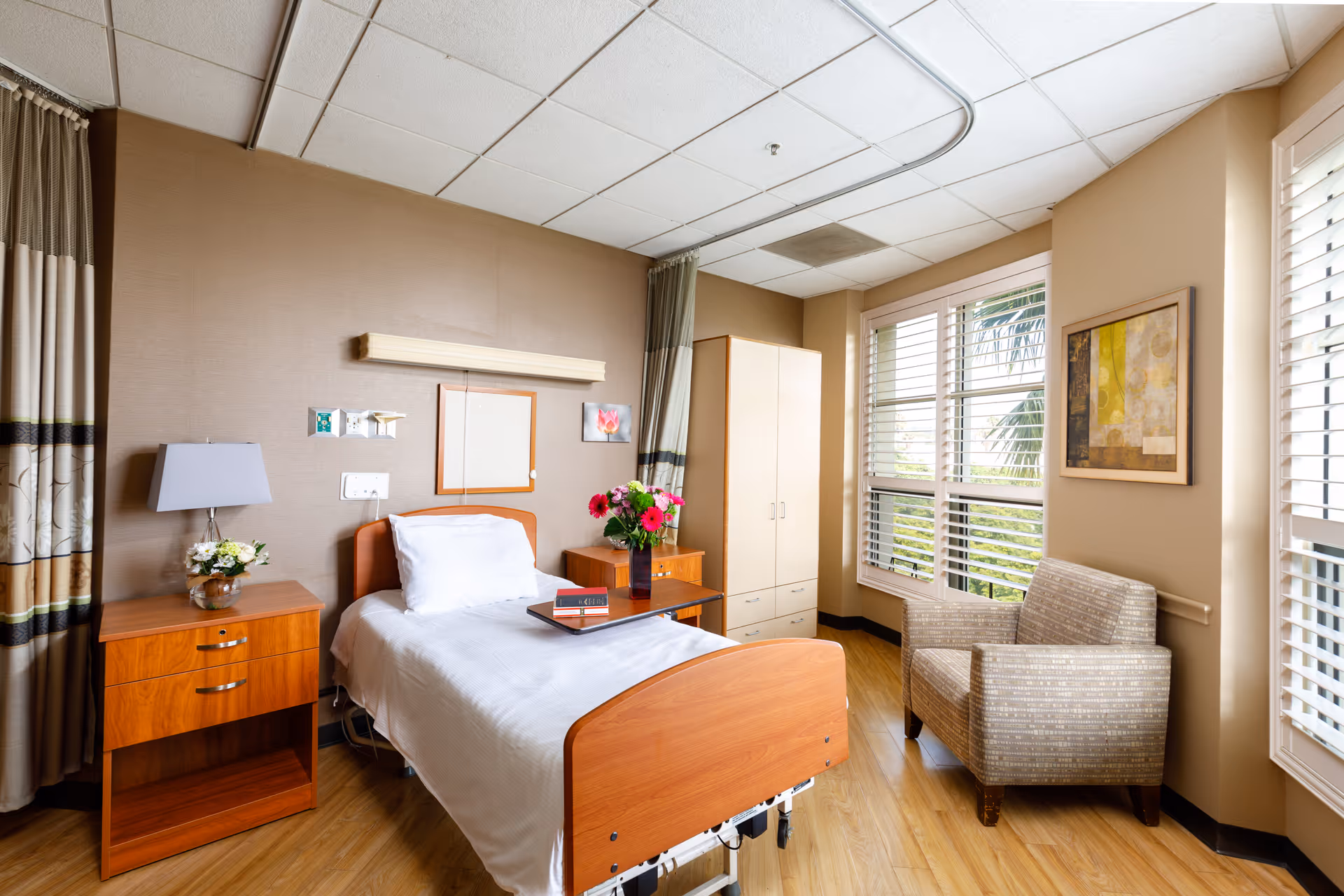A bright and clean healthcare facility bedroom with a single hospital bed, white linens, a wooden headboard and footboard, a bedside table with a lamp and flowers, a small cabinet with a vase of flowers, a comfortable armchair, large windows with white blinds, and a framed abstract painting on the wall.