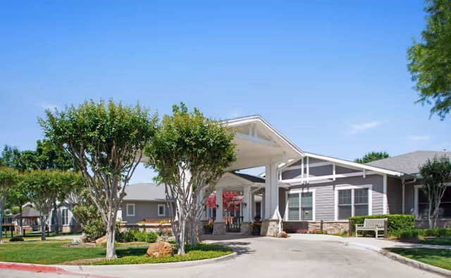 Exterior view of a single-story senior living facility building with a covered entrance driveway, surrounded by neatly trimmed trees, green lawns, and clear blue sky.