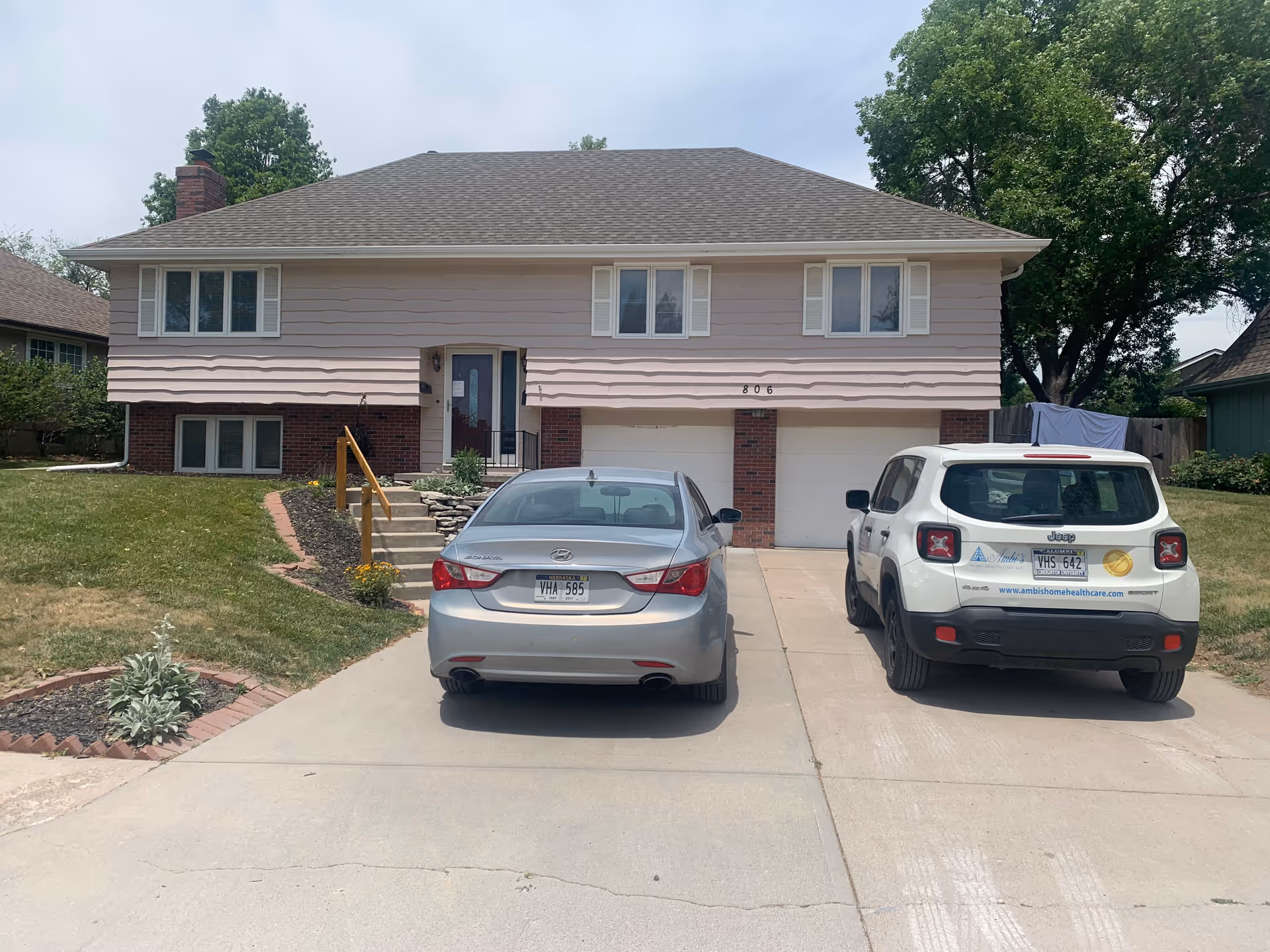 Two-story beige house with brick foundation, front stairs, and two cars parked in driveway.