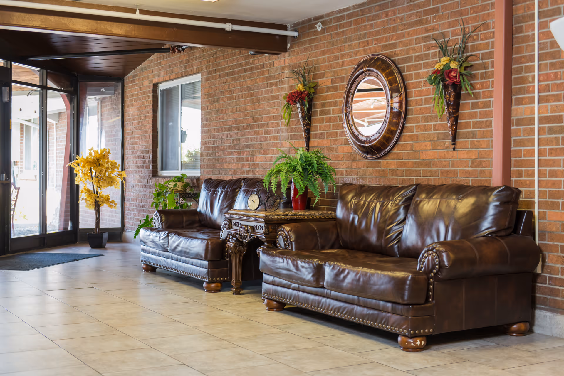 A cozy seating area in a facility with two brown leather sofas separated by an ornate wooden side table holding a clock and a potted fern. The background features a brick wall with a round decorative mirror and two wall-mounted floral arrangements. To the left, there is a glass door entrance with a yellow potted plant nearby.