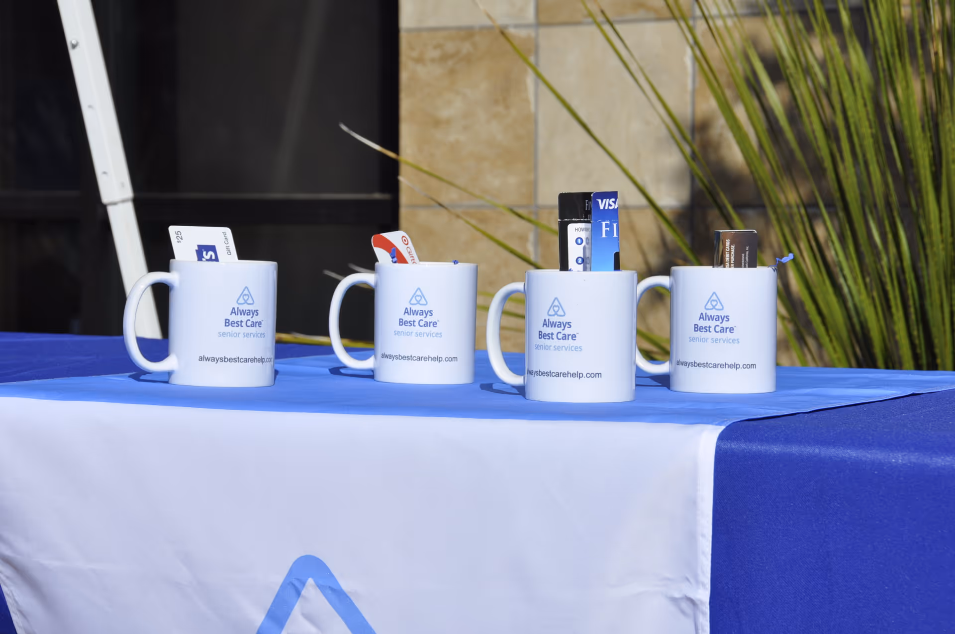 Four white mugs with Always Best Care logos on a blue outdoor table.