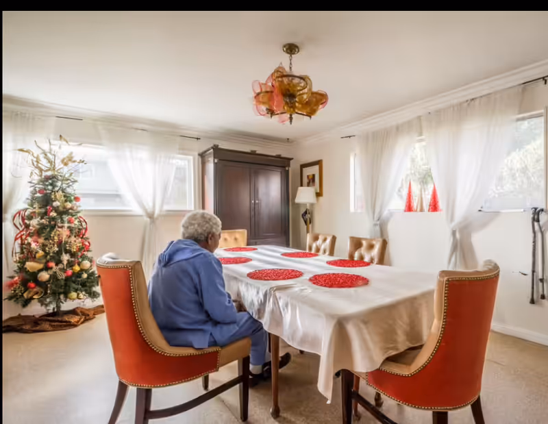 An elderly resident sits at a long dining table in a festive dining room.