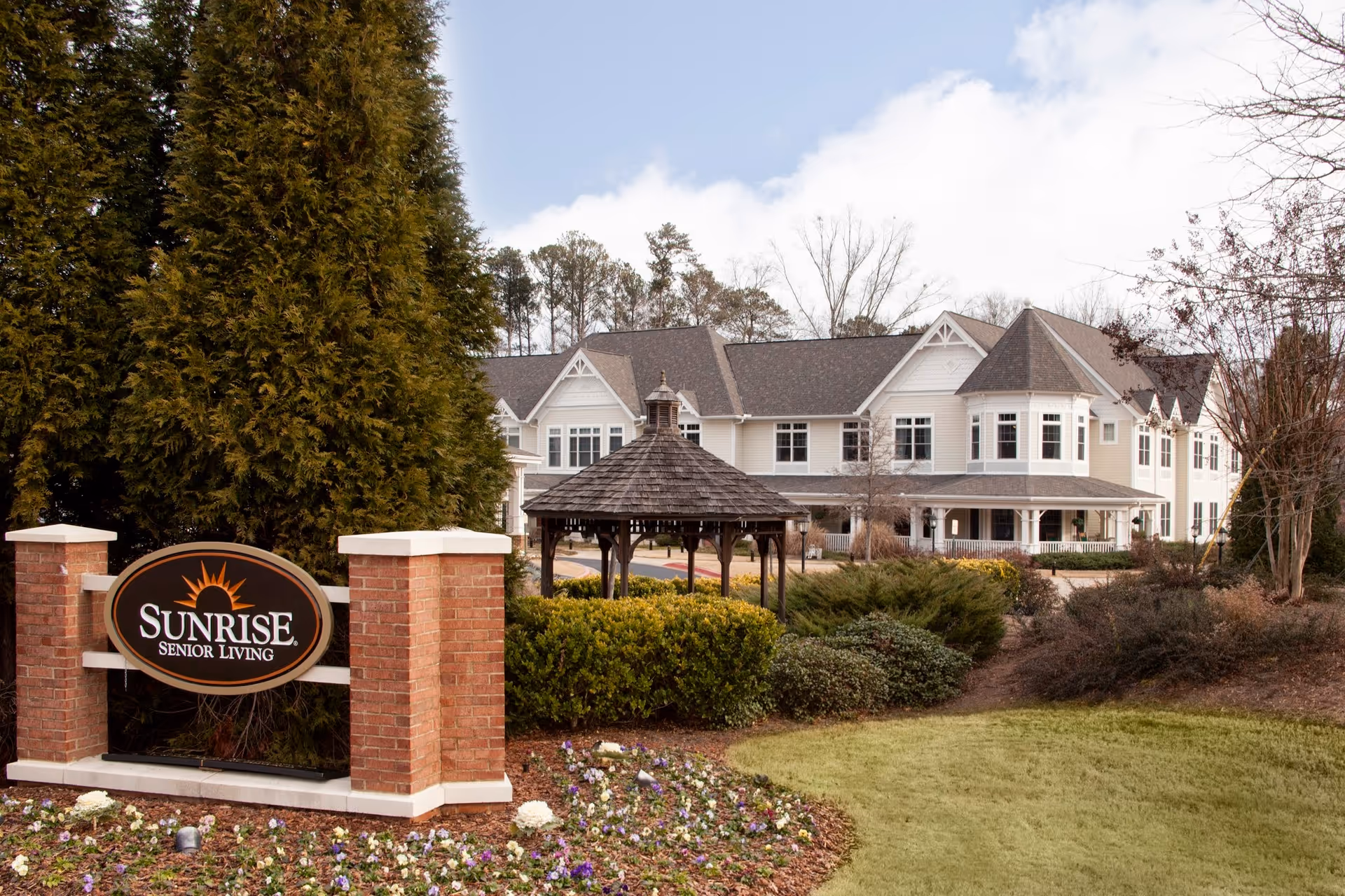 Exterior view of a senior living facility with a large two-story building in the background, surrounded by trees and landscaping. In the foreground, there is a brick sign with the logo and text 'Sunrise Senior Living' and a wooden gazebo behind it.