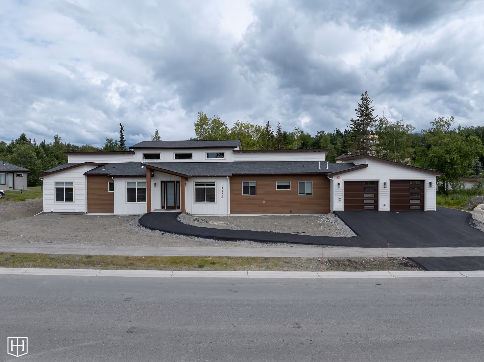 Front exterior view of a modern single-story building with a combination of white and brown siding, large windows, a double garage with dark brown doors, and a newly paved driveway. The building is surrounded by trees and under a cloudy sky.
