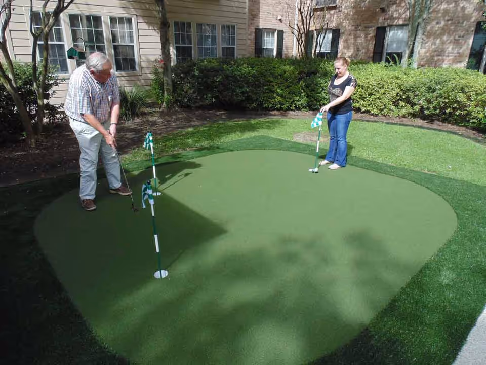 An elderly man and a woman playing mini golf on a small artificial putting green outside a residential building with bushes and windows in the background.