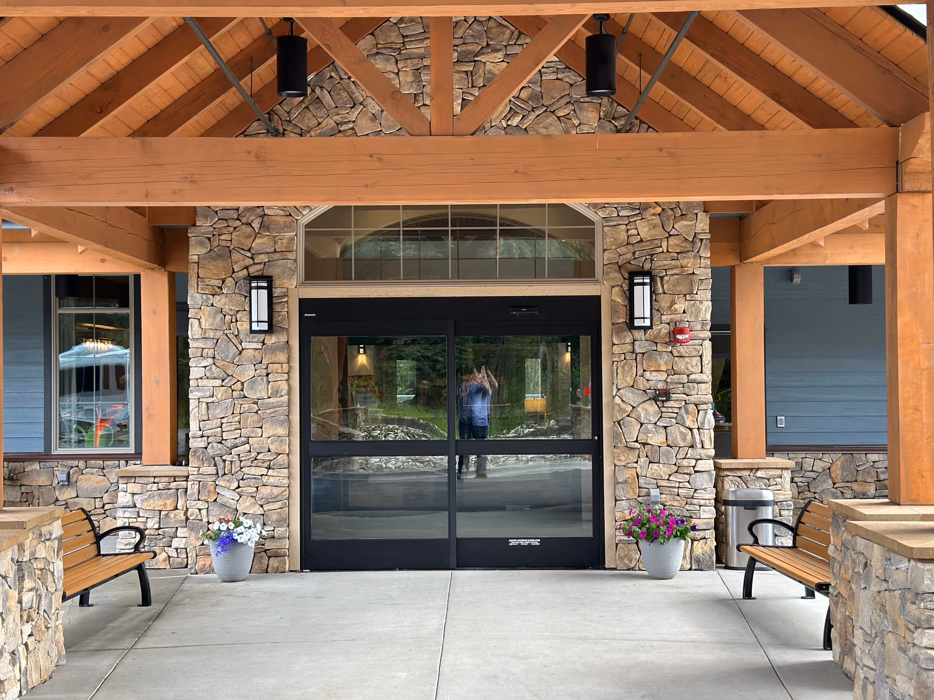 Timber Creek Senior Living entrance with stone walls, wood beams, glass doors, benches, and planters.