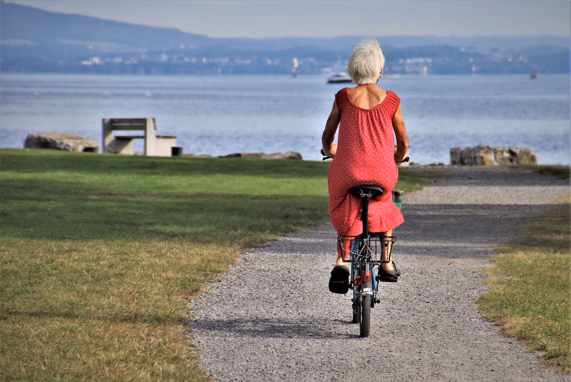 An elderly woman rides a bicycle along a gravel path beside a lake.