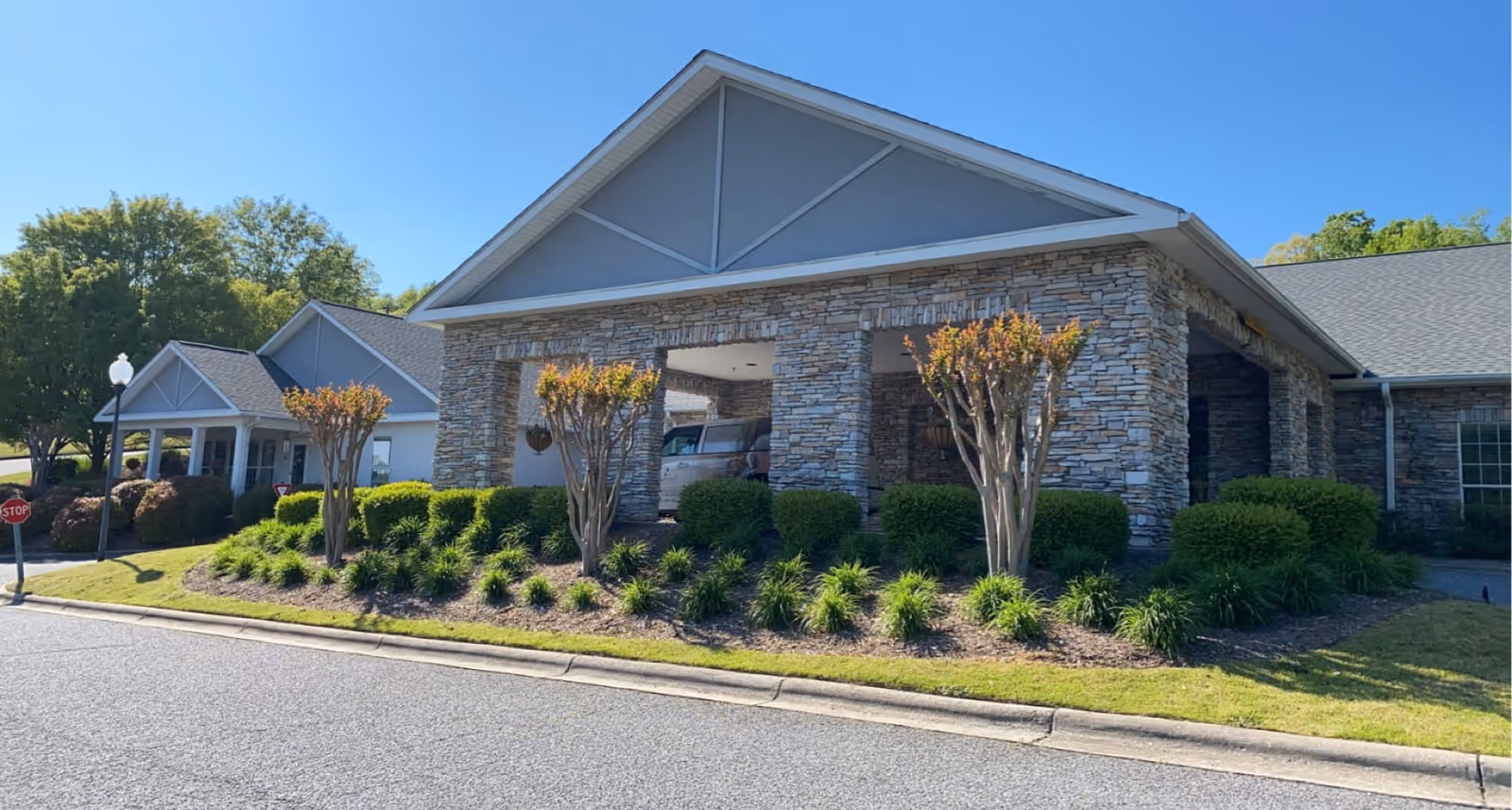 Exterior view of a senior living facility building with stone and siding facade, a covered entrance, neatly trimmed bushes, small trees, and a clear blue sky.