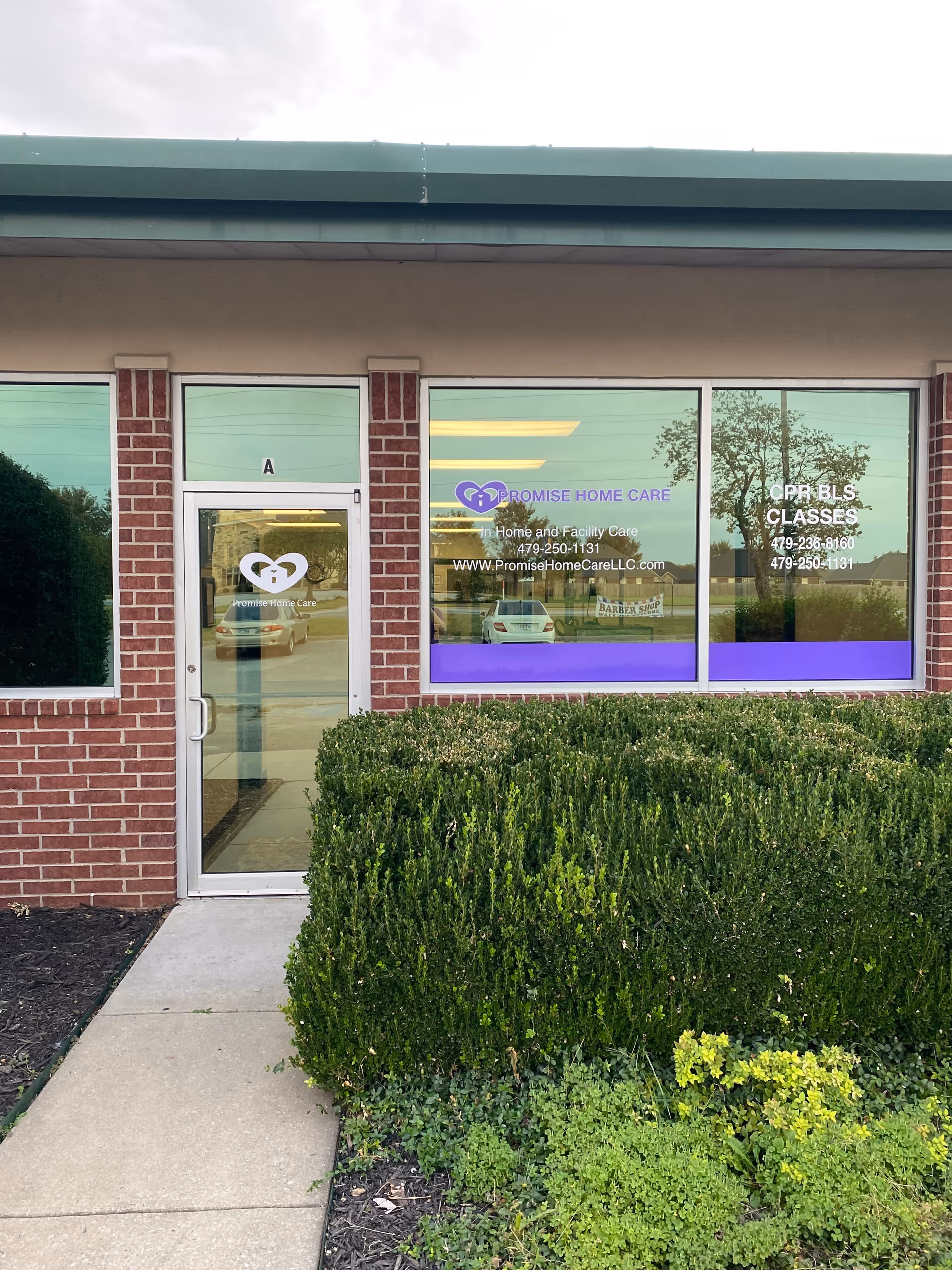 Brick storefront with glass doors and windows, Promise Home Care signage, trimmed hedges outside.