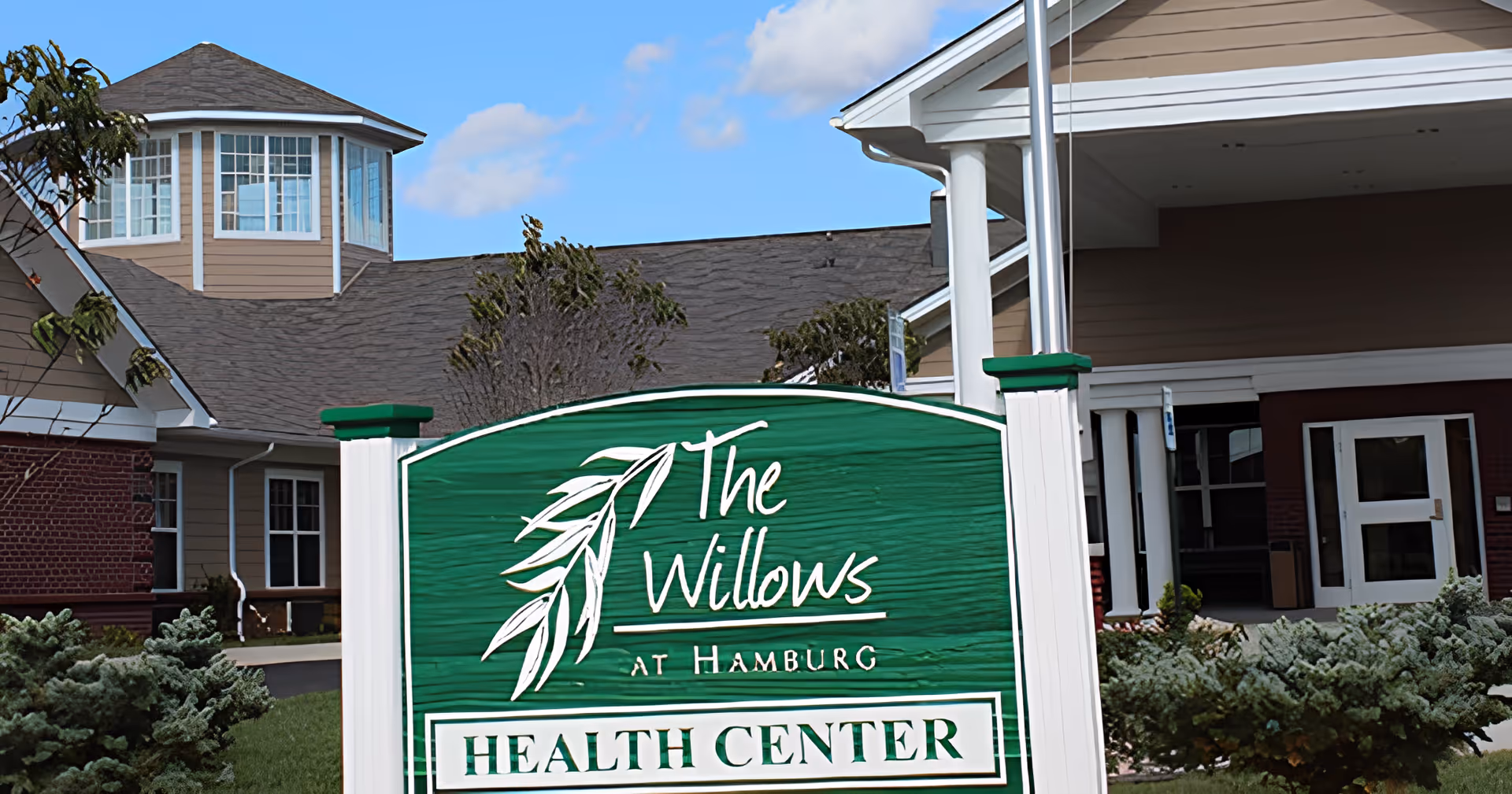Exterior view of The Willows At Hamburg Health Center building with a green sign in the foreground displaying the facility's name. The building has a beige and brick facade with white trim and a turret with multiple windows under a blue sky with some clouds.